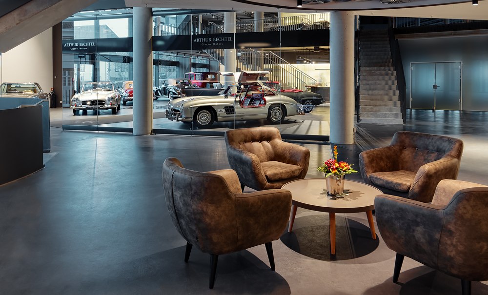 Moderne Lobby mit vier Sesseln um einen Tisch, Blumenarrangement. Im Hintergrund sind Oldtimer hinter Glas ausgestellt., © V8 HOTEL Motorworld Region Stuttgart Moderne Lobby mit vier Sesseln um einen Tisch, Blumenarrangement. Im Hintergrund sind Oldtimer hinter Glas ausgestellt., © V8 HOTEL Motorworld Region Stuttgart