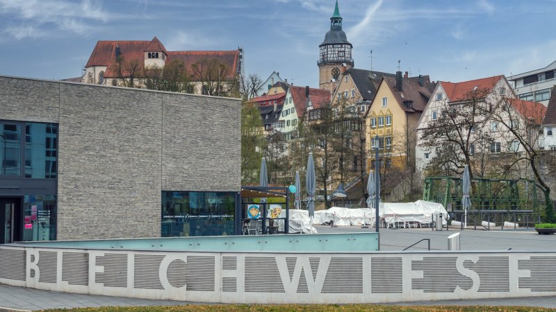 Stadtansicht von Backnang mit modernen und historischen Gebäuden, im Vordergrund der Schriftzug 'Bleichwiese' und ein Platz mit geschlossenen Sonnenschirmen., © René Straube