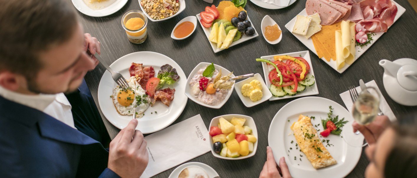Ein Tisch mit einem vielfältigen Frühstücksbuffet: Eier, Aufschnitt, Obst, Müsli und Getränke. Zwei Personen sitzen am Tisch und essen., © ARCOTEL Hotels