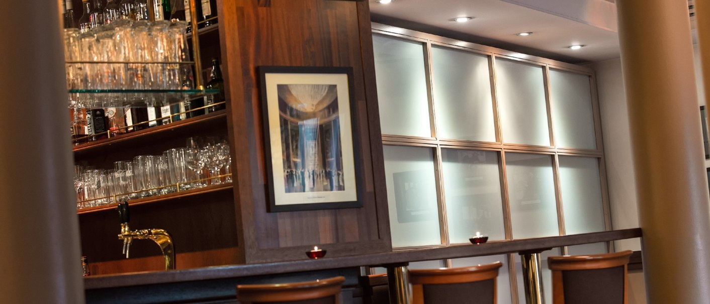 Stilvolle Bar mit Holztheke, Barhockern und beleuchteten Regalen voller Gläser und Flaschen. Dezente Beleuchtung und ein Bild an der Wand., © TOMAS