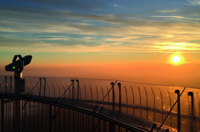 Aussichtsplattform eines Fernsehturms bei Sonnenaufgang. Ein Fernrohr und ein Glasgeländer sind sichtbar, während die Sonne am Horizont aufgeht., © SWR Media Services GmbH / Fernsehturm Stuttgart