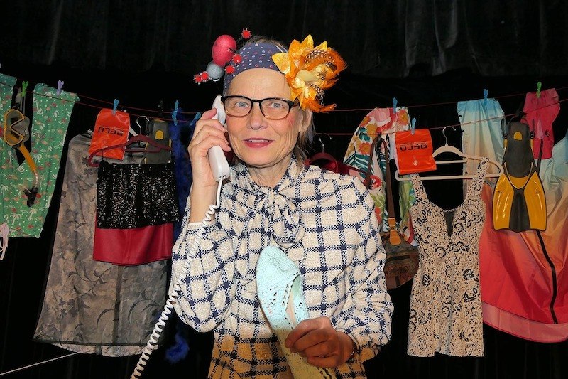 Eine Frau mit einem auff&auml;lligen Blumenhut telefoniert. Im Hintergrund h&auml;ngt eine W&auml;scheleine mit bunten Kleidungsst&uuml;cken und Taucherzubeh&ouml;r., &copy; Galli Theater Backnang