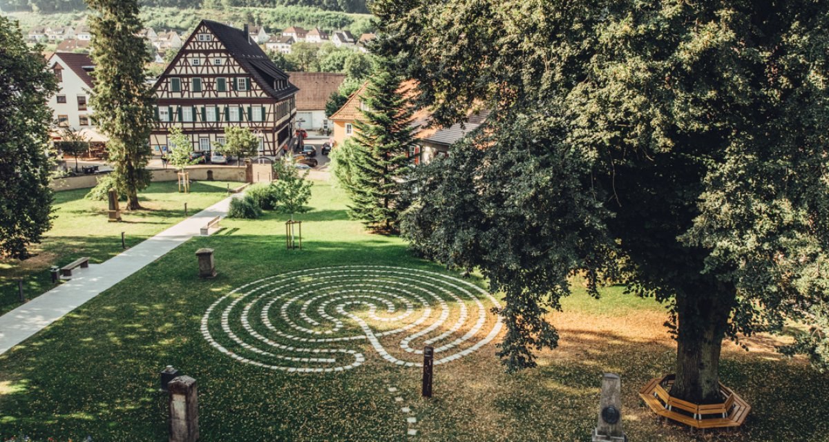 Ein Labyrinth aus Steinen im Kirchgarten, umgeben von Bäumen und einem Fachwerkhaus im Hintergrund. Ein ruhiger, grüner Ort., © Tourismus Ostalb Ein Labyrinth aus Steinen im Kirchgarten, umgeben von Bäumen und einem Fachwerkhaus im Hintergrund. Ein ruhiger, grüner Ort., © Tourismus Ostalb