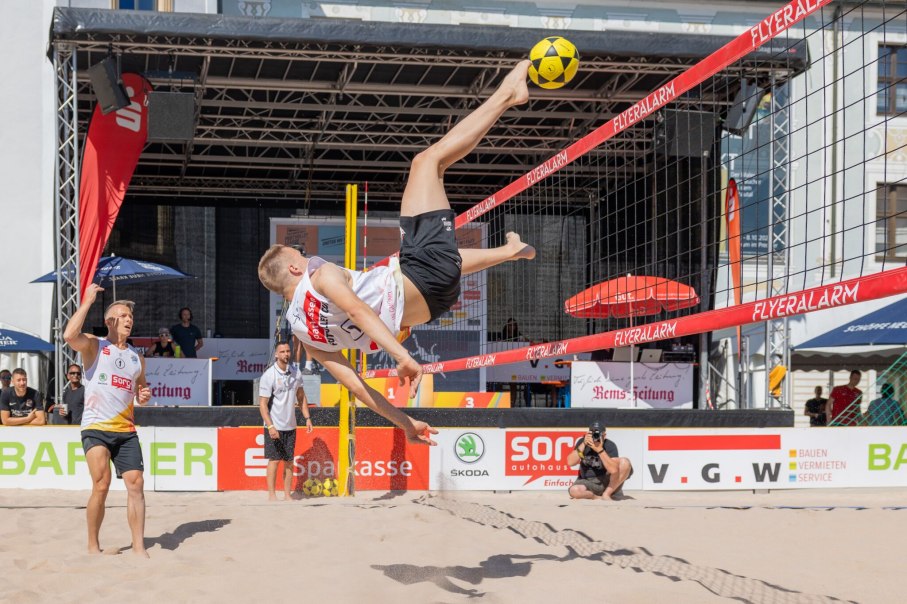 Ein Beachvolleyballspieler f&uuml;hrt einen spektakul&auml;ren Fallr&uuml;ckzieher aus, w&auml;hrend sein Teamkollege zuschaut. Im Hintergrund sind Zuschauer und Werbebanner sichtbar., &copy; F&uuml;r die Bildrechte: Amt f&uuml;r Medien und Kommunikation Schw&auml;bisch Gm&uuml;nd