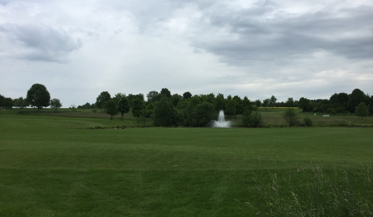 Weitläufiger Golfplatz mit grünen Wiesen, Bäumen und einem Springbrunnen. Der Himmel ist bewölkt, was eine ruhige Atmosphäre schafft., © www.pro-cycl.de