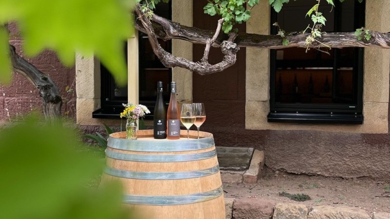 Ein Weinfass dient als Tisch mit zwei Weinflaschen und Gl&auml;sern darauf, umgeben von Weinreben vor einem rustikalen Geb&auml;ude., &copy; Weingut Schwarz