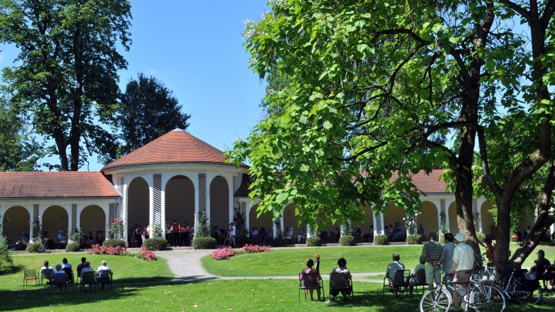 Menschen genie&szlig;en einen sonnigen Tag im Kurpark Bad Boll. Sie sitzen und stehen vor einem Pavillon mit roten Dachziegeln, umgeben von gr&uuml;nen B&auml;umen., &copy; Gemeindearchiv Bad Boll