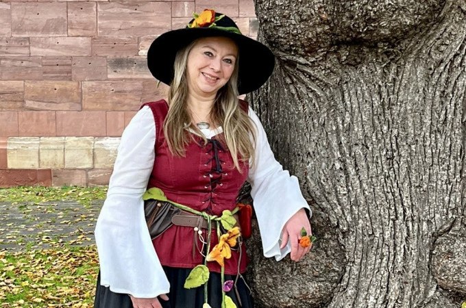 Eine Frau in mittelalterlicher Kleidung mit Blumen am Gürtel lehnt lächelnd an einem großen Baum vor einer Steinmauer., © Stadtverwaltung Weil der Stadt