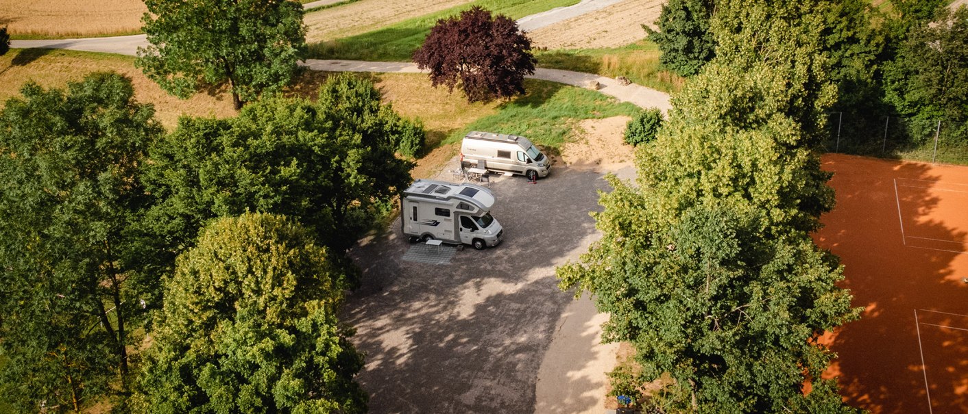 Zwei Wohnmobile stehen auf einem Stellplatz, umgeben von Bäumen. Ein Tennisplatz ist rechts im Bild zu sehen., © Blu Dolci Artwork