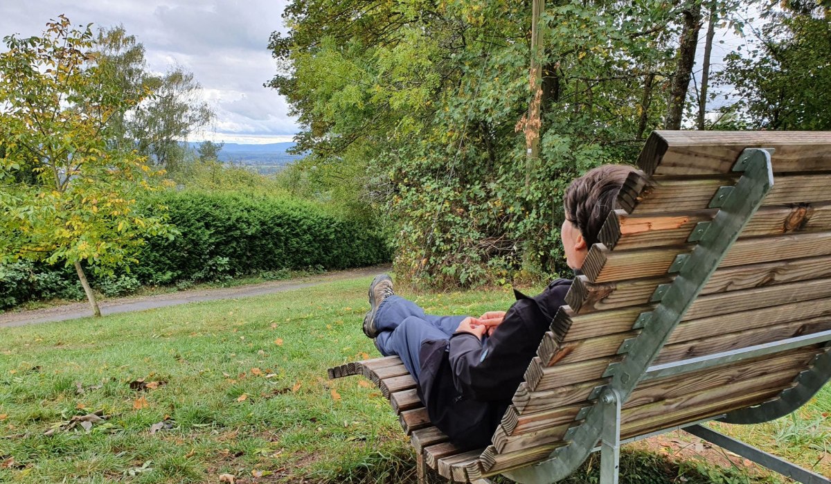 Eine Person sitzt entspannt auf einer Holzbank und blickt auf eine grüne, bewaldete Landschaft mit bewölktem Himmel. Eine Person sitzt entspannt auf einer Holzbank und blickt auf eine grüne, bewaldete Landschaft mit bewölktem Himmel.