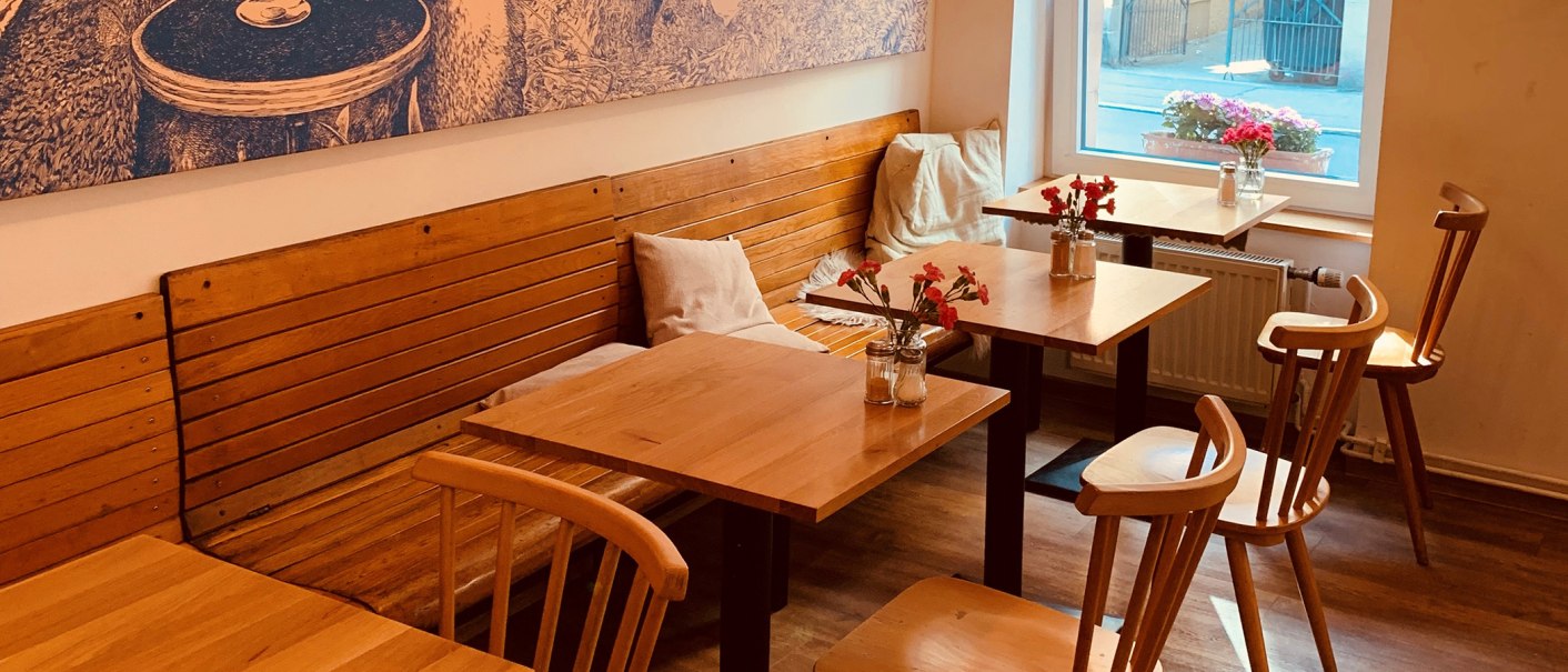 Gemütlicher Innenbereich eines Cafés mit Holztischen und -stühlen, Blumenvasen auf den Tischen und einem großen Fenster mit Blick nach draußen., © Gustav Gastro GmbH