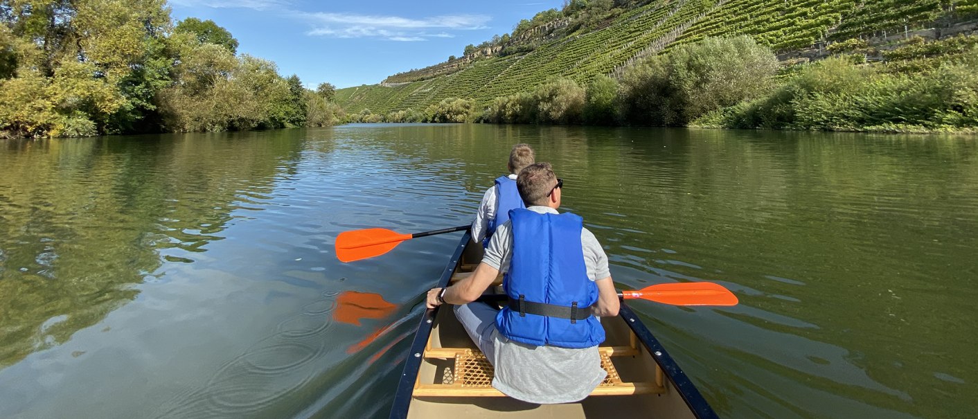 Zwei Personen paddeln in einem Kanu auf einem ruhigen Fluss, umgeben von grünen Bäumen und Weinbergen unter blauem Himmel., © Matthias Pflüger - Rock-the-River.com Zwei Personen paddeln in einem Kanu auf einem ruhigen Fluss, umgeben von grünen Bäumen und Weinbergen unter blauem Himmel., © Matthias Pflüger - Rock-the-River.com