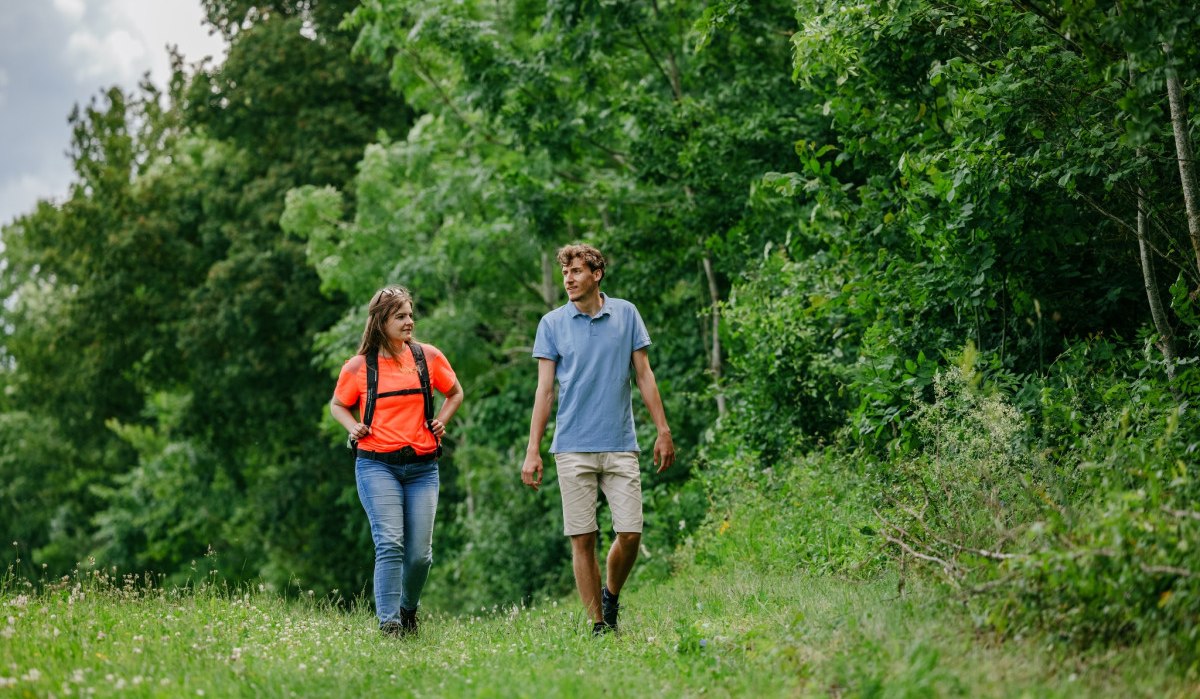 Zwei Personen wandern auf einem grünen Pfad, umgeben von dichtem Wald. Eine Person trägt ein orangefarbenes Shirt, die andere ein blaues., © Landkreis Göppingen Zwei Personen wandern auf einem grünen Pfad, umgeben von dichtem Wald. Eine Person trägt ein orangefarbenes Shirt, die andere ein blaues., © Landkreis Göppingen