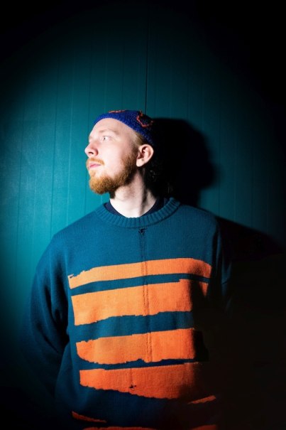 Ein Mann in einem blau-orange gestreiften Pullover steht vor einem dunklen Hintergrund und blickt zur Seite., &copy; Kulturhaus Schwanen