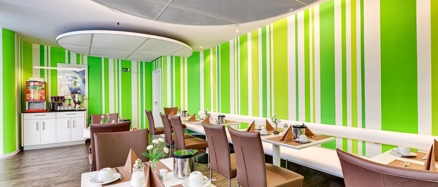 Moderner Frühstücksraum mit grünen Streifenwänden, gedeckten Tischen und braunen Stühlen. Ein Getränkeautomat steht in der Ecke., © attimo Hotel Stuttgart