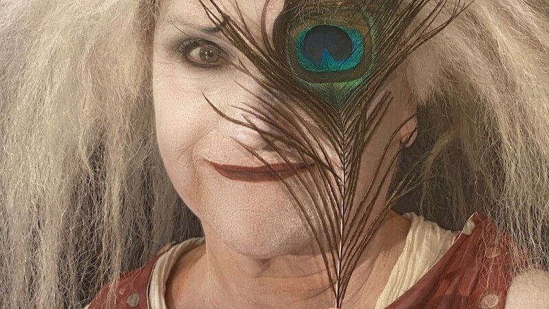 Eine Person mit wildem, weißem Haar und weiß geschminktem Gesicht hält eine Pfauenfeder vor einem Auge. Sie trägt ein rotes, gepunktetes Oberteil., © Mauerwerk GmbH