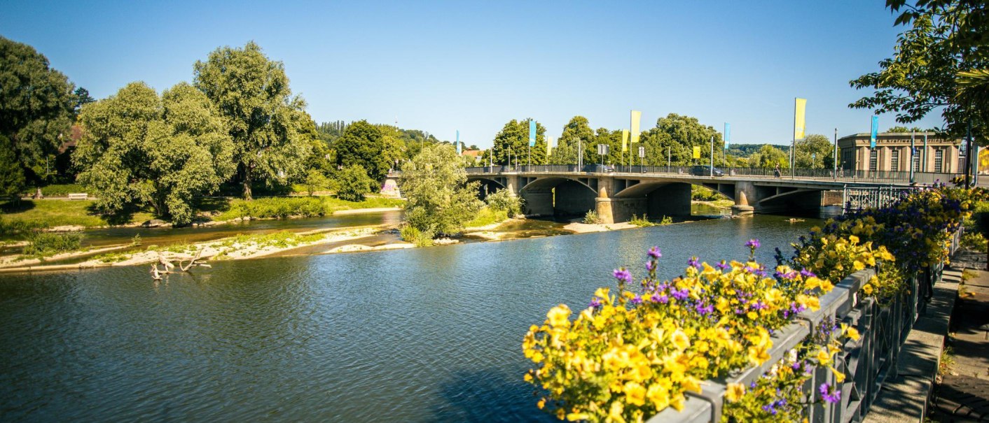 Sommerliche Flusslandschaft in Nürtingen mit einer Brücke und blühenden Blumen im Vordergrund. Der Himmel ist klar und blau., © Stuttgart-Marketing GmbH, Sarah Schmid Sommerliche Flusslandschaft in Nürtingen mit einer Brücke und blühenden Blumen im Vordergrund. Der Himmel ist klar und blau., © Stuttgart-Marketing GmbH, Sarah Schmid