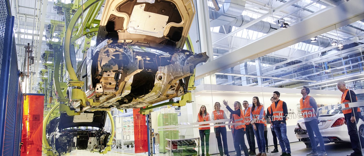 Besucher in orangefarbenen Westen beobachten die Autoproduktion im Mercedes-Benz Kundencenter. Ein Fahrzeugchassis hängt an einem Förderband., © Mercedes-Benz Kundencenter Besucher in orangefarbenen Westen beobachten die Autoproduktion im Mercedes-Benz Kundencenter. Ein Fahrzeugchassis hängt an einem Förderband., © Mercedes-Benz Kundencenter