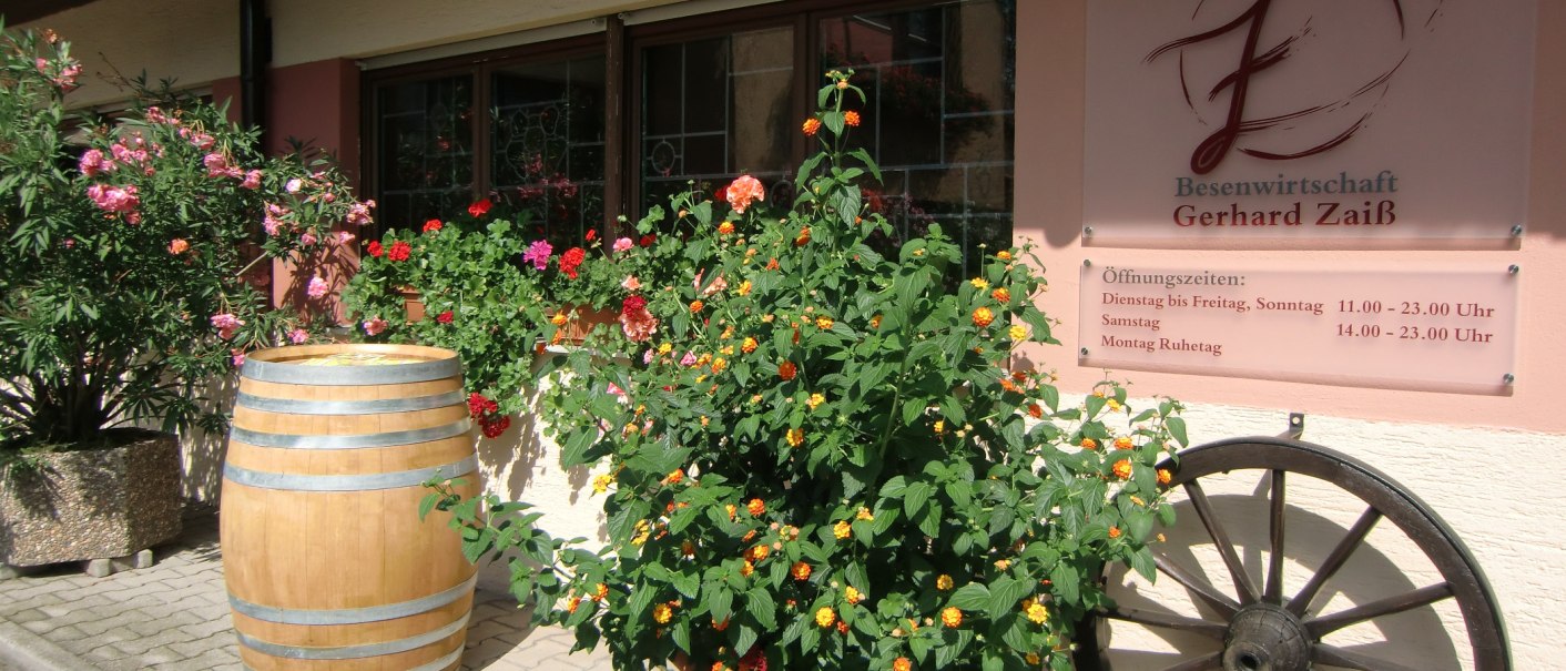 Eingang der Besenwirtschaft Gerhard Zaiß mit blühenden Pflanzen, einem Weinfass und einem Schild mit Öffnungszeiten., © Simone Mack Eingang der Besenwirtschaft Gerhard Zaiß mit blühenden Pflanzen, einem Weinfass und einem Schild mit Öffnungszeiten., © Simone Mack