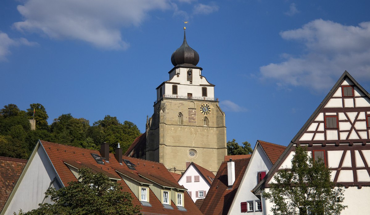 Die Stiftskirche Herrenberg erhebt sich hinter malerischen Fachwerkhäusern unter einem klaren blauen Himmel., © Natur.Nah. Schönbuch & Heckengäu Die Stiftskirche Herrenberg erhebt sich hinter malerischen Fachwerkhäusern unter einem klaren blauen Himmel., © Natur.Nah. Schönbuch & Heckengäu