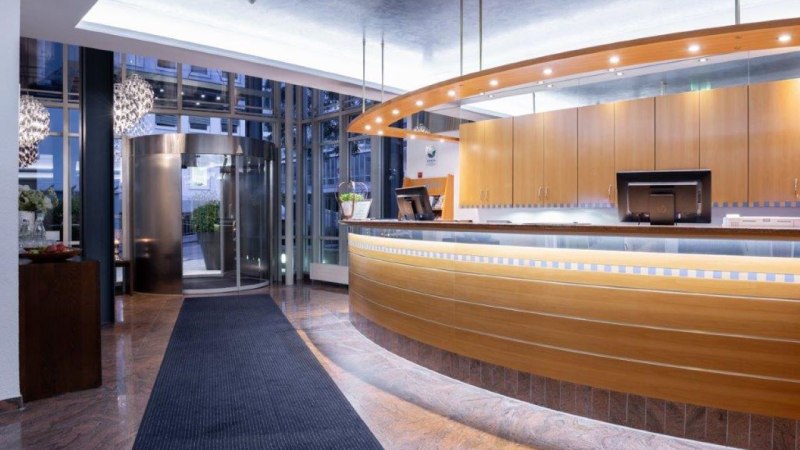 Moderne Hotelrezeption mit geschwungener Holztheke, beleuchtetem Empfangsbereich und Drehtür im Hintergrund. Elegantes, einladendes Ambiente., © Kronen Hotel GmbH Moderne Hotelrezeption mit geschwungener Holztheke, beleuchtetem Empfangsbereich und Drehtür im Hintergrund. Elegantes, einladendes Ambiente., © Kronen Hotel GmbH