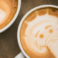Zwei Cappuccinos mit kunstvoller Latte Art: links ein Blattmuster, rechts ein Elefant, auf einem Holztisch stehend., © Marc Fuhrmann