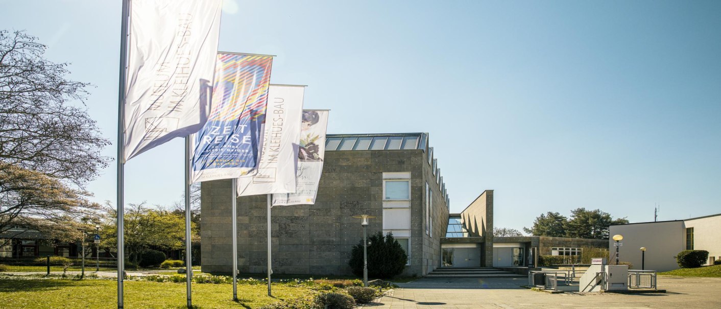 Das Museum Im Kleinhues-Bau in Kornwestheim, moderne Architektur mit mehreren Fahnen im Vordergrund, sonniger Tag., © SMG, Sarah Schmid Das Museum Im Kleinhues-Bau in Kornwestheim, moderne Architektur mit mehreren Fahnen im Vordergrund, sonniger Tag., © SMG, Sarah Schmid