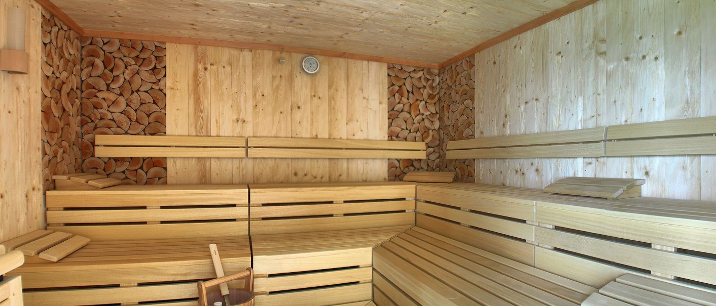 Innenansicht einer Sauna aus hellem Holz mit Bänken, einem Holzeimer und gestapeltem Holz an den Wänden., © TOMAS