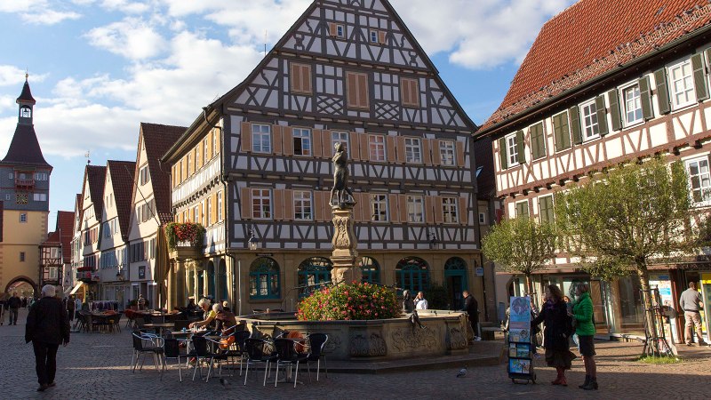 Marktplatz in Winnenden mit Fachwerkhäusern, einem zentralen Brunnen und Menschen, die den Platz beleben. Im Hintergrund ein Turm mit Uhr., © Stuttgart-Marketing GmbH