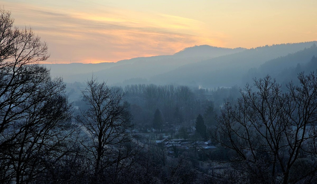 Sonnenuntergang über nebliger Landschaft, Bäume im Vordergrund, Hügel im Hintergrund. Der Himmel ist in warmen Farben gefärbt. Sonnenuntergang über nebliger Landschaft, Bäume im Vordergrund, Hügel im Hintergrund. Der Himmel ist in warmen Farben gefärbt.