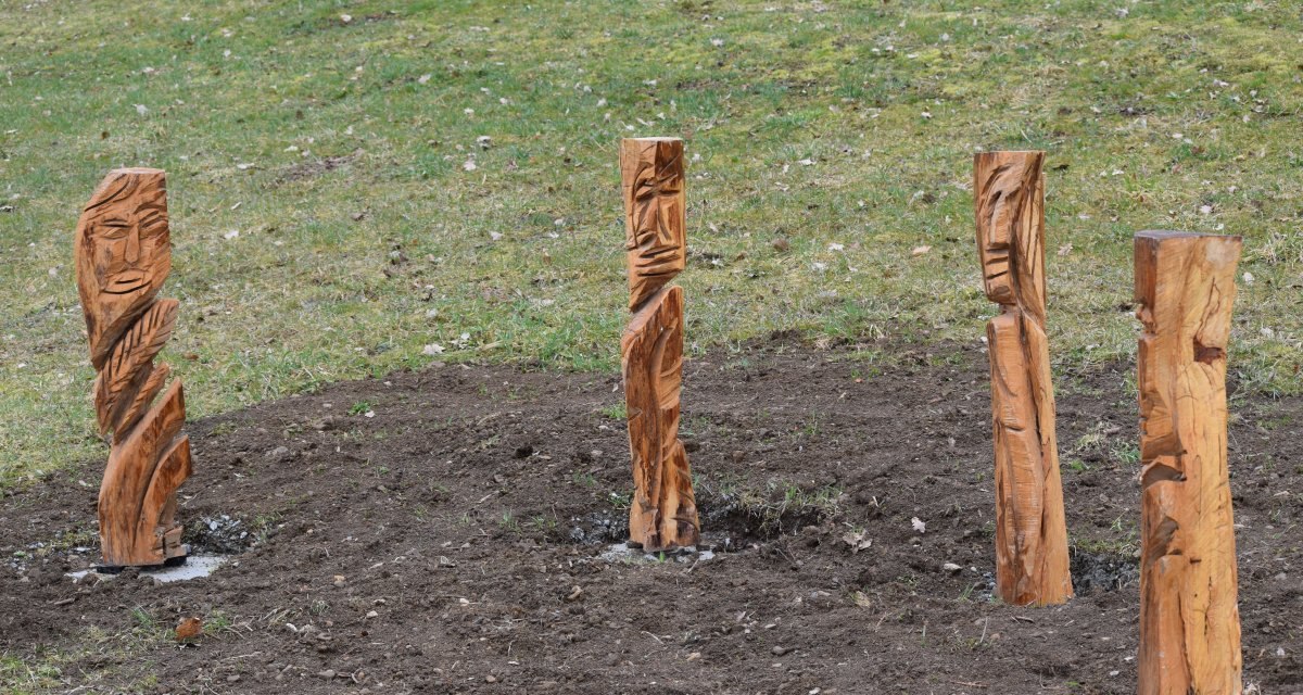 Vier geschnitzte Holzskulpturen stehen auf einem Erdstreifen, umgeben von Gras. Die Skulpturen zeigen abstrakte Gesichter., &copy; Gemeinde B&ouml;bingen