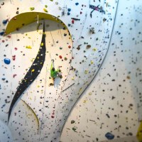 Ein Kletterer in grüner Kleidung erklimmt eine hohe Indoor-Kletterwand mit bunten Griffen im DAV Kletter- und Boulderzentrum Schwaben., © Rockerei