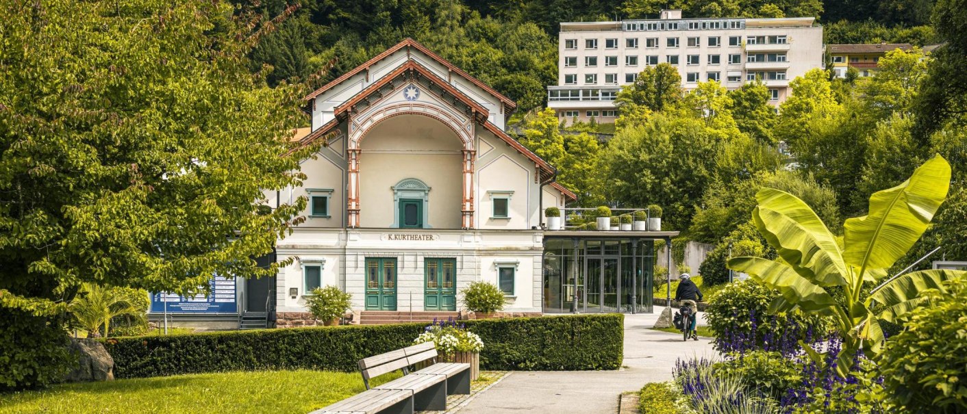 Das Kurtheater steht inmitten eines grünen Gartens mit Bäumen und Pflanzen. Ein Weg führt vorbei, auf dem eine Person mit Fahrrad zu sehen ist., © Stuttgart-Marketing GmbH, Sarah Schmid
