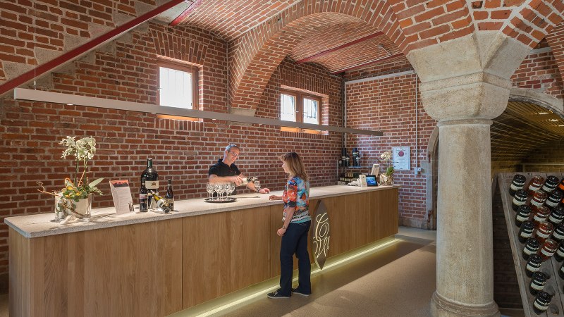 Eine Theke in einer Weinkellerei mit Backsteinwänden. Ein Mann schenkt einer Frau Wein ein. Weinflaschen sind auf der Theke ausgestellt., © Collegium Wirtemberg