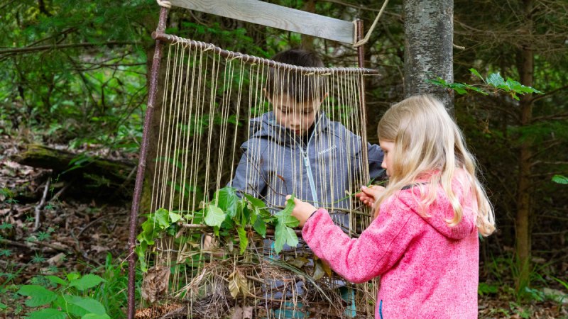 Zwei Kinder weben Blätter und Zweige in einen großen Holzrahmen im Wald. Sie sind konzentriert und umgeben von Bäumen und Pflanzen., © Touristikgemeinschaft Hohenlohe, Künzelsau / Stadtverwaltung Künzelsau