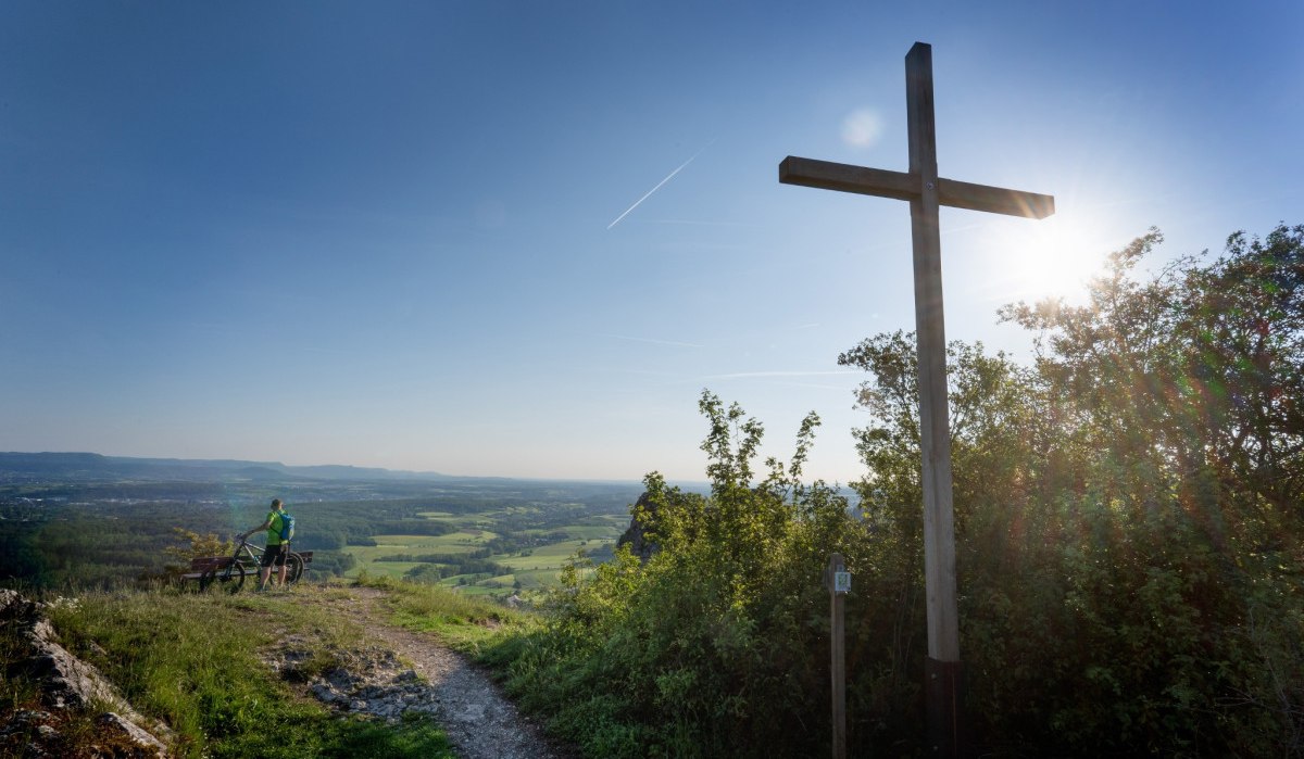Ein Gipfelkreuz auf einem Hügel, daneben ein Radfahrer mit Fahrrad, der die weite Landschaft betrachtet. Die Sonne scheint hell am Himmel., © Landkreis Göppingen