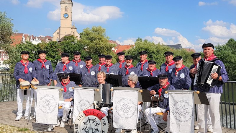 Eine Männergruppe in Matrosenuniformen mit Instrumenten posiert vor einer Kirche. Im Hintergrund sind Bäume und ein Kirchturm zu sehen., © Neckar-Knurrhähne