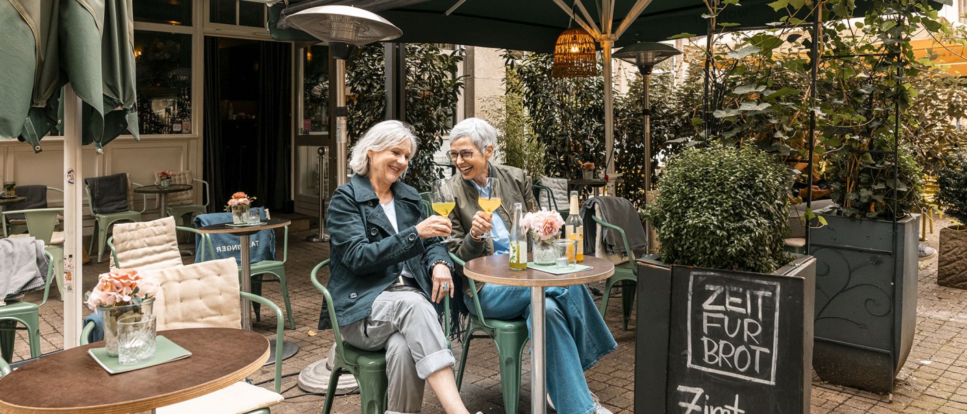 Zwei Frauen sitzen in einem gemütlichen Café im Freien, stoßen mit Getränken an und lachen. Ein Schild bietet Zimtschnecken an., © Stuttgart Marketing GmbH - Sarah Schmid Zwei Frauen sitzen in einem gemütlichen Café im Freien, stoßen mit Getränken an und lachen. Ein Schild bietet Zimtschnecken an., © Stuttgart Marketing GmbH - Sarah Schmid