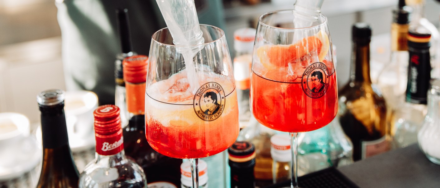 Ein Barkeeper füllt zwei Gläser mit einem Getränk an einer Bar. Im Hintergrund sind Flaschen und Gläser zu sehen., © L. Prestle/ Documenting Love Ein Barkeeper füllt zwei Gläser mit einem Getränk an einer Bar. Im Hintergrund sind Flaschen und Gläser zu sehen., © L. Prestle/ Documenting Love