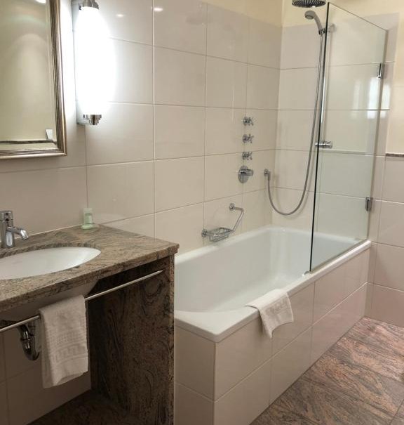 Modernes Badezimmer mit Badewanne, Duschkopf, Glasabtrennung und Waschbecken mit Spiegel. Helle Fliesen und Handtuchhalter sind sichtbar., © Hotel Royal Modernes Badezimmer mit Badewanne, Duschkopf, Glasabtrennung und Waschbecken mit Spiegel. Helle Fliesen und Handtuchhalter sind sichtbar., © Hotel Royal