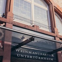 Fassade der Weinmanufaktur Untertürkheim mit Glasvordach, Schriftzug und dekorativen Fenstern., © Weinmanufaktur Stuttgart Fassade der Weinmanufaktur Untertürkheim mit Glasvordach, Schriftzug und dekorativen Fenstern., © Weinmanufaktur Stuttgart