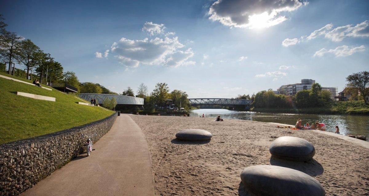 Sonniger Tag am Flussufer mit Sandstrand, großen Steinen und einer Brücke im Hintergrund. Menschen entspannen am Wasser., © Stoppel Sonniger Tag am Flussufer mit Sandstrand, großen Steinen und einer Brücke im Hintergrund. Menschen entspannen am Wasser., © Stoppel