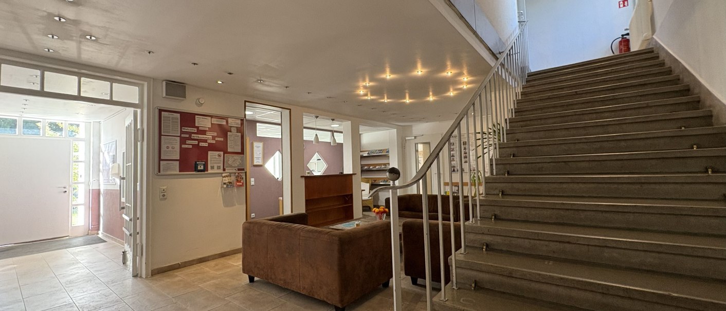 Heller Eingangsbereich mit Treppe, braunen Sofas und Infotafel. Moderne Beleuchtung und Fliesenboden schaffen eine einladende Atmosphäre., © TOMAS Heller Eingangsbereich mit Treppe, braunen Sofas und Infotafel. Moderne Beleuchtung und Fliesenboden schaffen eine einladende Atmosphäre., © TOMAS
