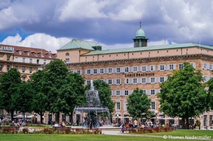Ein belebter Platz in Stuttgart mit einem Brunnen und dem Gebäude "Carls Brauhaus" im Hintergrund, umgeben von Bäumen und Menschen., © Stuttgart Marketing GmbH Ein belebter Platz in Stuttgart mit einem Brunnen und dem Gebäude "Carls Brauhaus" im Hintergrund, umgeben von Bäumen und Menschen., © Stuttgart Marketing GmbH