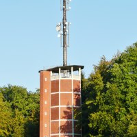 Roter Backstein-Wasserturm mit Antennenmast, umgeben von grünen Bäumen unter klarem Himmel.