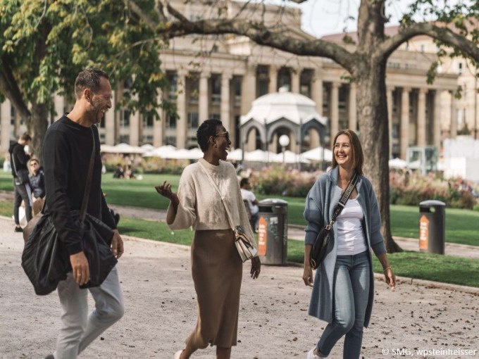 Drei Personen spazieren l&auml;chelnd durch einen Park in Stuttgart. Im Hintergrund sind B&auml;ume und ein historisches Geb&auml;ude zu sehen., &copy; Stuttgart Marketing GmbH