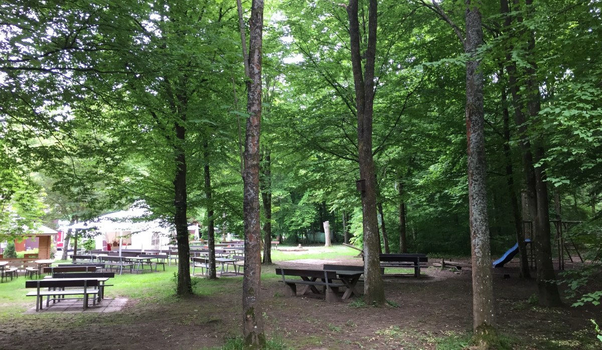 Ein ruhiger Biergarten im Wald mit leeren Holztischen und Bänken unter grünen Bäumen. Im Hintergrund sind Zelte und ein Spielplatz zu sehen., © www.pro-cycl.de