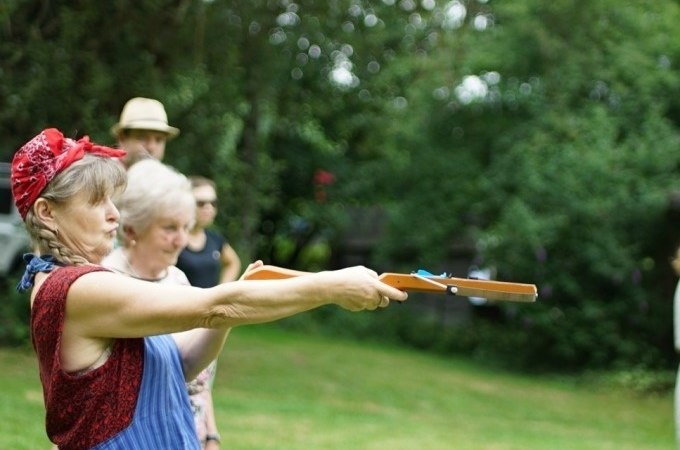 Eine Frau zielt mit einem Spielzeuggewehr bei einem Outdoor-Event. Weitere Personen stehen im Hintergrund auf einer Wiese., &copy; Cool-Tours StattReisen
