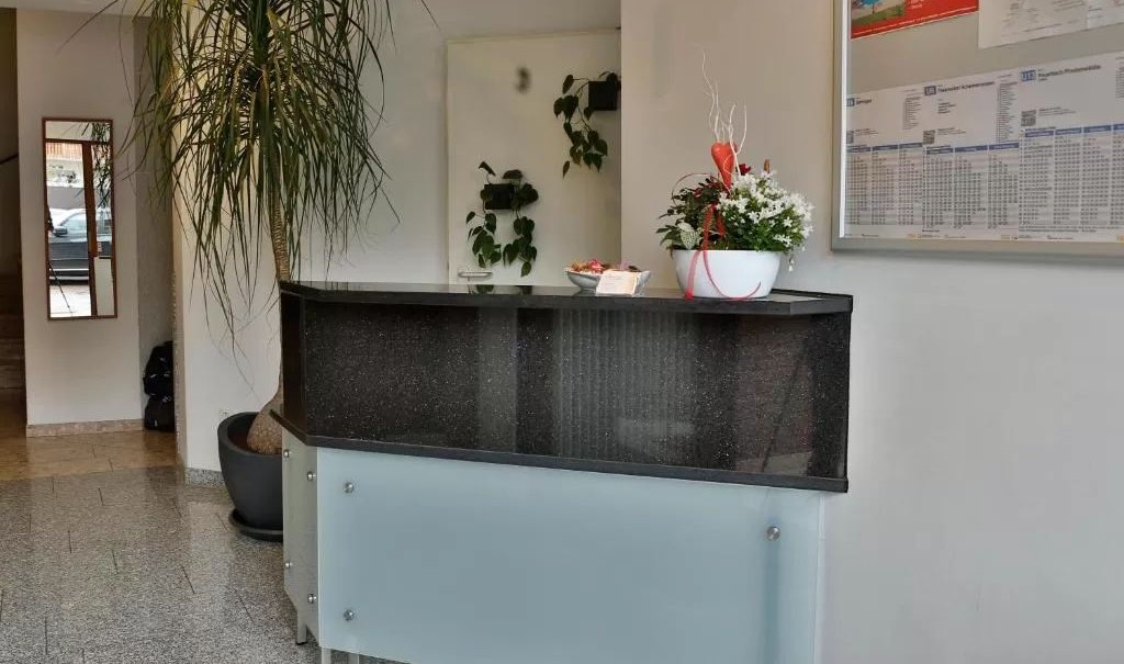 Ein moderner Rezeptionstresen mit einer Pflanze und Dekoration. An der Wand hängt ein Informationsbrett. Der Boden ist gefliest., © Feuerbacher Hof Ein moderner Rezeptionstresen mit einer Pflanze und Dekoration. An der Wand hängt ein Informationsbrett. Der Boden ist gefliest., © Feuerbacher Hof