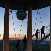 Menschen steigen eine Wendeltreppe im Schönbuchturm hinauf, umgeben von Holzsäulen. Im Hintergrund ist ein Sonnenuntergang über einer weiten Landschaft zu sehen., © Stuttgart-Marketing GmbH, Martina Denker